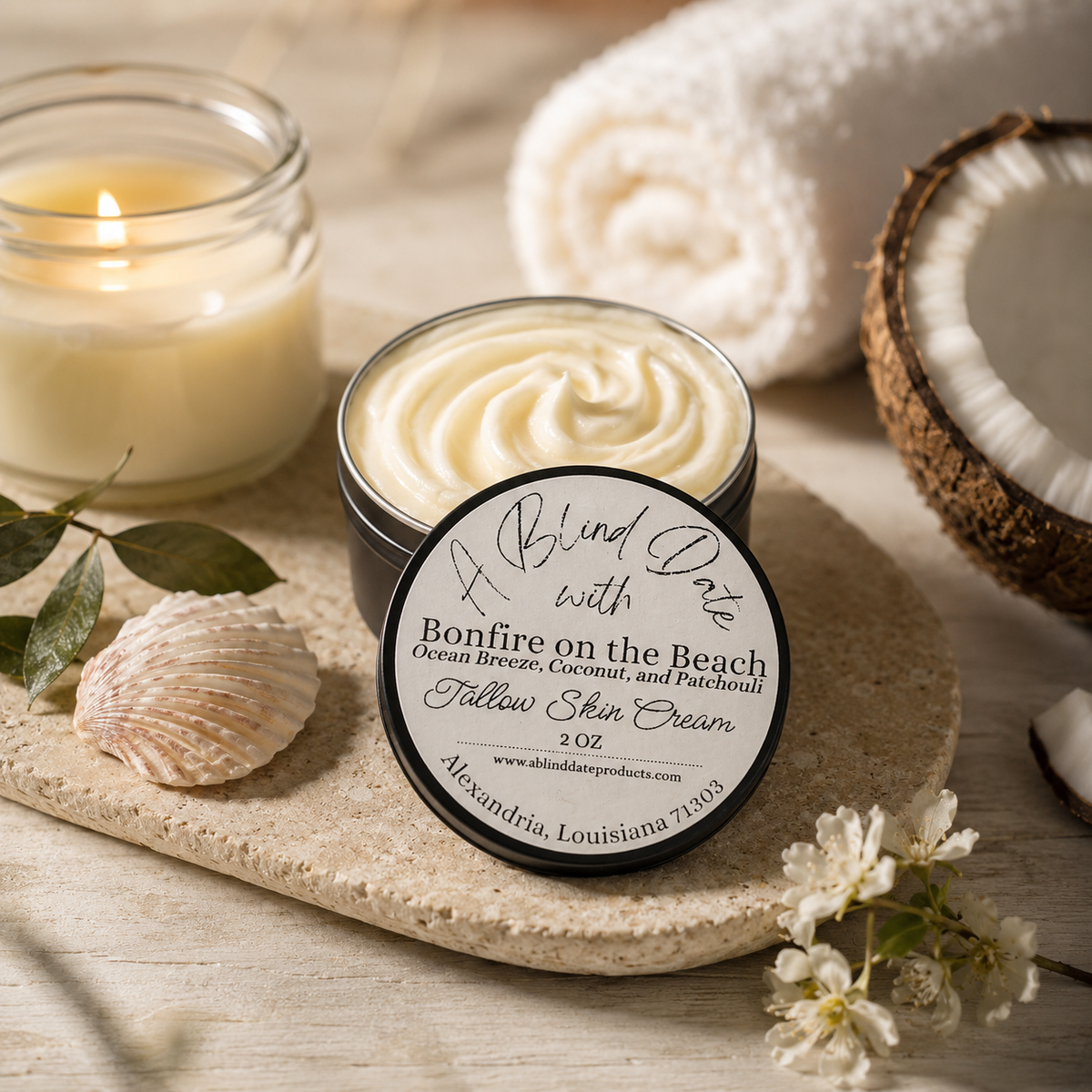 Open jar of Skindria 'Bonfire on the Beach' Follow Skin Cream with its lid leaning in front, styled on a stone tray with a lit candle, rolled towel, coconut halves, seashell, and small white flowers.
