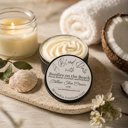 Open jar of Skindria 'Bonfire on the Beach' Follow Skin Cream with its lid leaning in front, styled on a stone tray with a lit candle, rolled towel, coconut halves, seashell, and small white flowers.