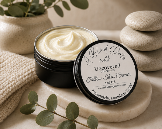 An open black jar of creamy yellow-toned body cream sits beside its lid on a stone slab, with eucalyptus leaves and stacked stones in a spa-style setting. The lid label reads 'Bind Date with Uncovered Towel Skin Cream' and includes the website www.